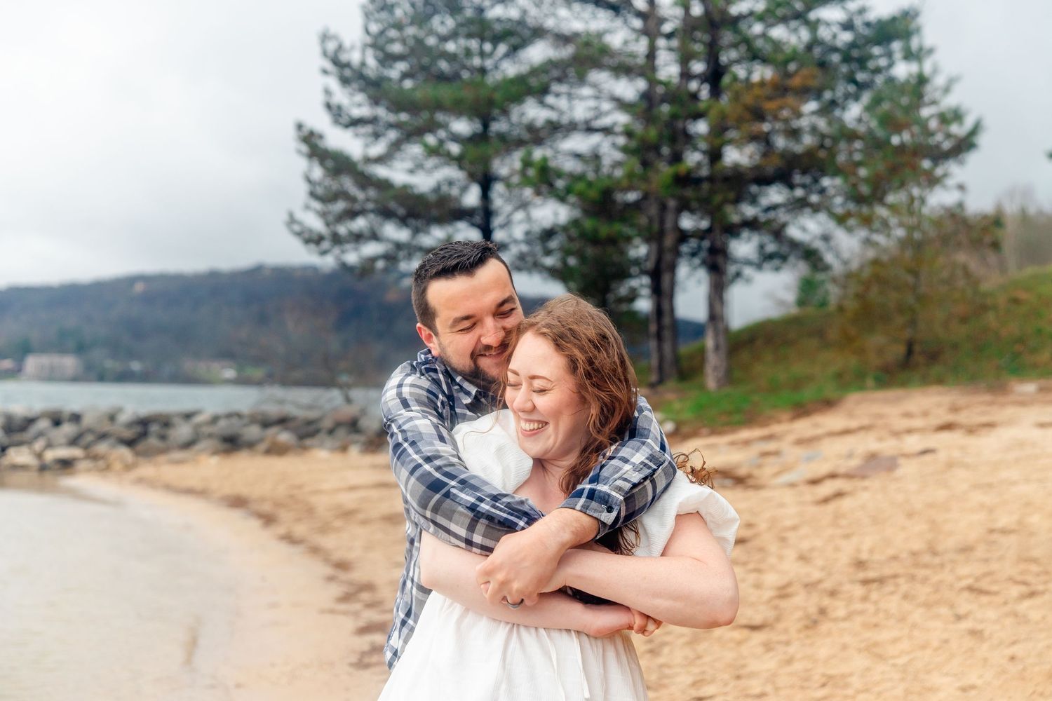 Couple embracing and laughing on a sandy beach with pine trees and hills in the background. deep creek lake stat park engagement photos