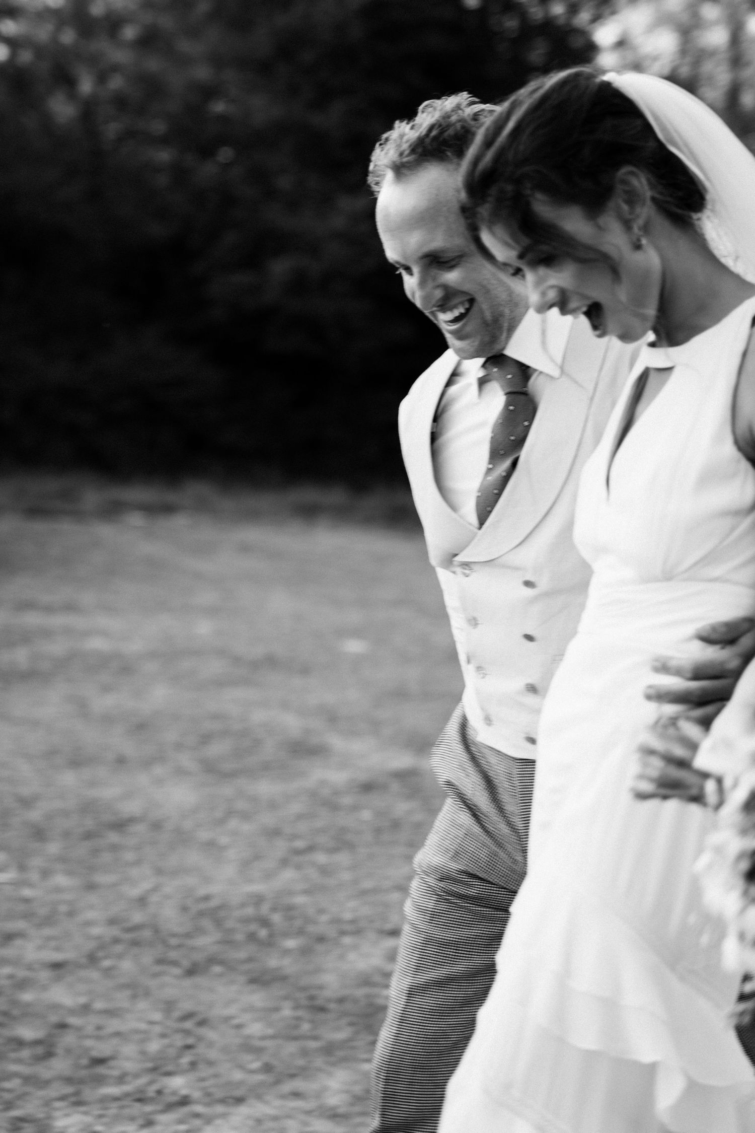 Black and white photo of a laughing bride and groom walking together outdoors on their wedding day.