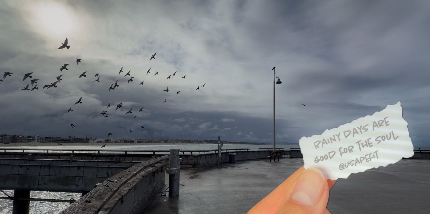 A hand holds a note about rainy days near a pier with birds flying over stormy waters.