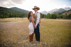 Couple posing in a mountain meadow, woman in white dress and boots, man in cowboy hat and jeans.