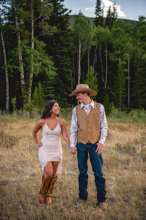 Couple holding hands in a mountain meadow, woman in white dress and cowboy boots, man in vest and cowboy hat.