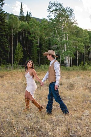 Couple in western attire holding hands in a golden meadow with pine trees, woman in white dress and boots, man in cowboy hat.