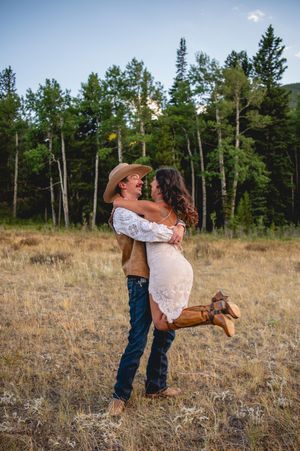 A man in a cowboy hat lifts a woman in a white lace dress and boots in a golden meadow with pine trees.