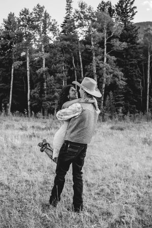 Black and white photo of a couple embracing in a meadow, man in cowboy hat lifting woman, surrounded by tall trees.
