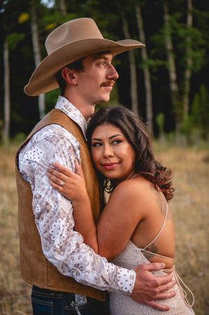 A couple in western attire embraces outdoors, man wearing cowboy hat and vest, woman in lace dress, with trees behind.