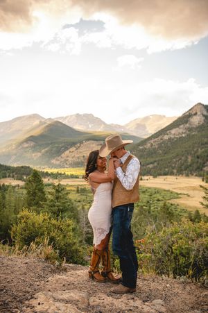Couple embracing on a rocky overlook with stunning Rocky Mountain backdrop, woman in white lace dress and cowboy boots, man in cowboy hat.