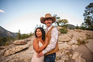 Couple posing on rocky terrain with mountain backdrop, woman in pink dress embracing man in cowboy hat and vest.