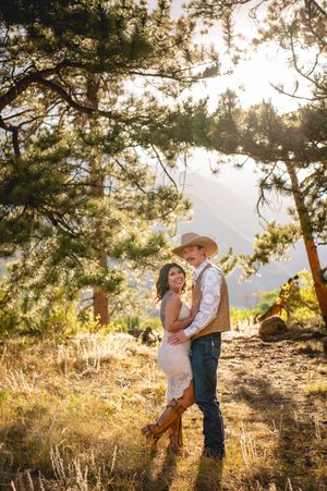 Couple embracing in golden sunlight among pine trees, woman in lace dress and boots, man in cowboy hat and jeans.