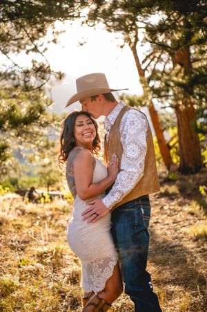 Couple embracing outdoors in golden sunlight, woman in lace dress smiling, man in cowboy hat and vest kissing her forehead.