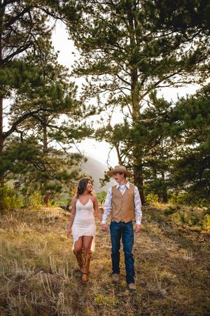 Couple walks hand-in-hand through pine forest, woman in white dress and cowboy boots, man in vest and cowboy hat.