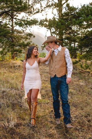 Couple in western attire walks through a meadow, woman in white lace dress and boots, man in cowboy hat and vest.