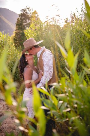 Couple kissing in a sunlit field surrounded by tall green plants, man wearing a tan cowboy hat and white lace shirt.