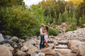 Couple embracing on rocky creek bed surrounded by lush green forest trees in a romantic outdoor setting.