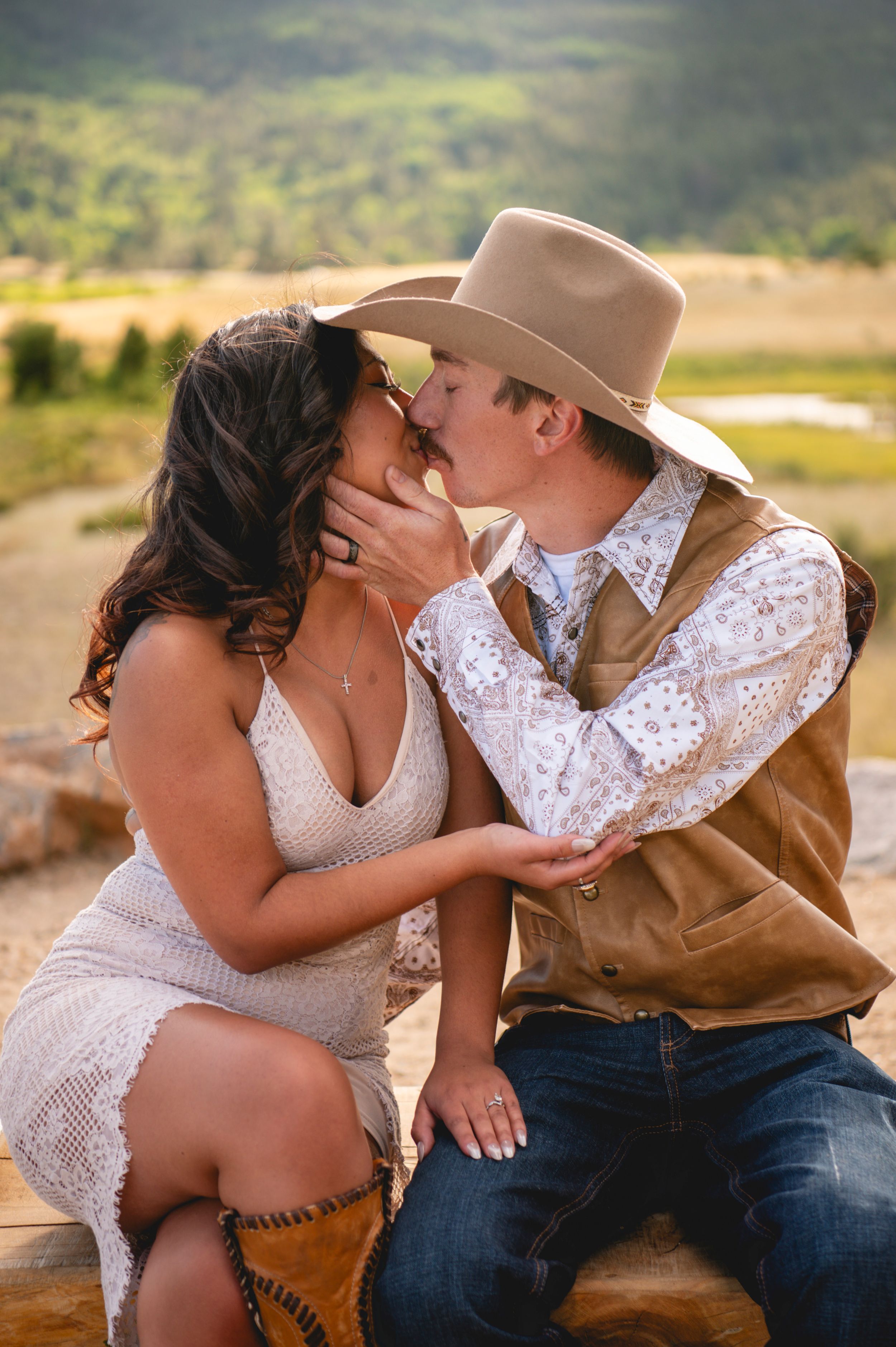 A couple shares a romantic kiss outdoors, woman in white lace dress and cowboy boots, man in tan vest and cowboy hat.