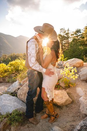 Couple kissing at golden hour on a rocky mountain overlook, woman in lace dress and cowboy boots, man in vest and hat.