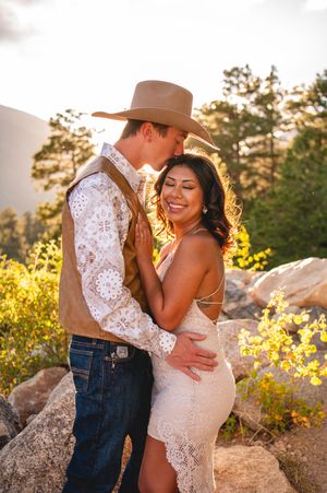 Couple embracing at golden hour in mountains, woman in lace dress, man in cowboy hat and vest, rocky outdoor setting.