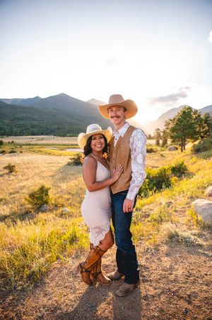 Couple in cowboy hats and western attire posing at golden hour with mountain meadow backdrop.