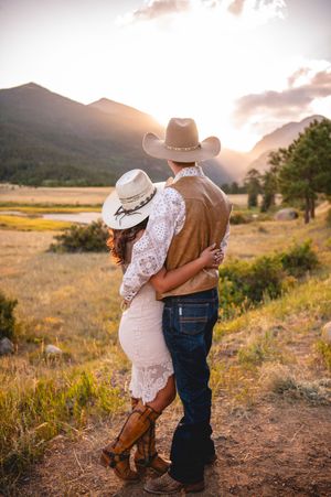 Couple in cowboy hats embracing at sunset in a scenic mountain meadow, wearing western attire including boots and jeans.