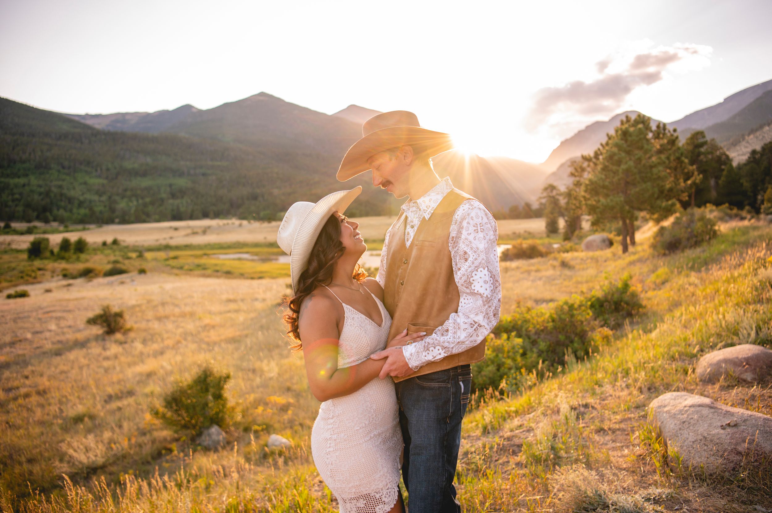Couple in cowboy hats embracing at golden sunset with mountain meadow backdrop in Rocky Mountain National Park.