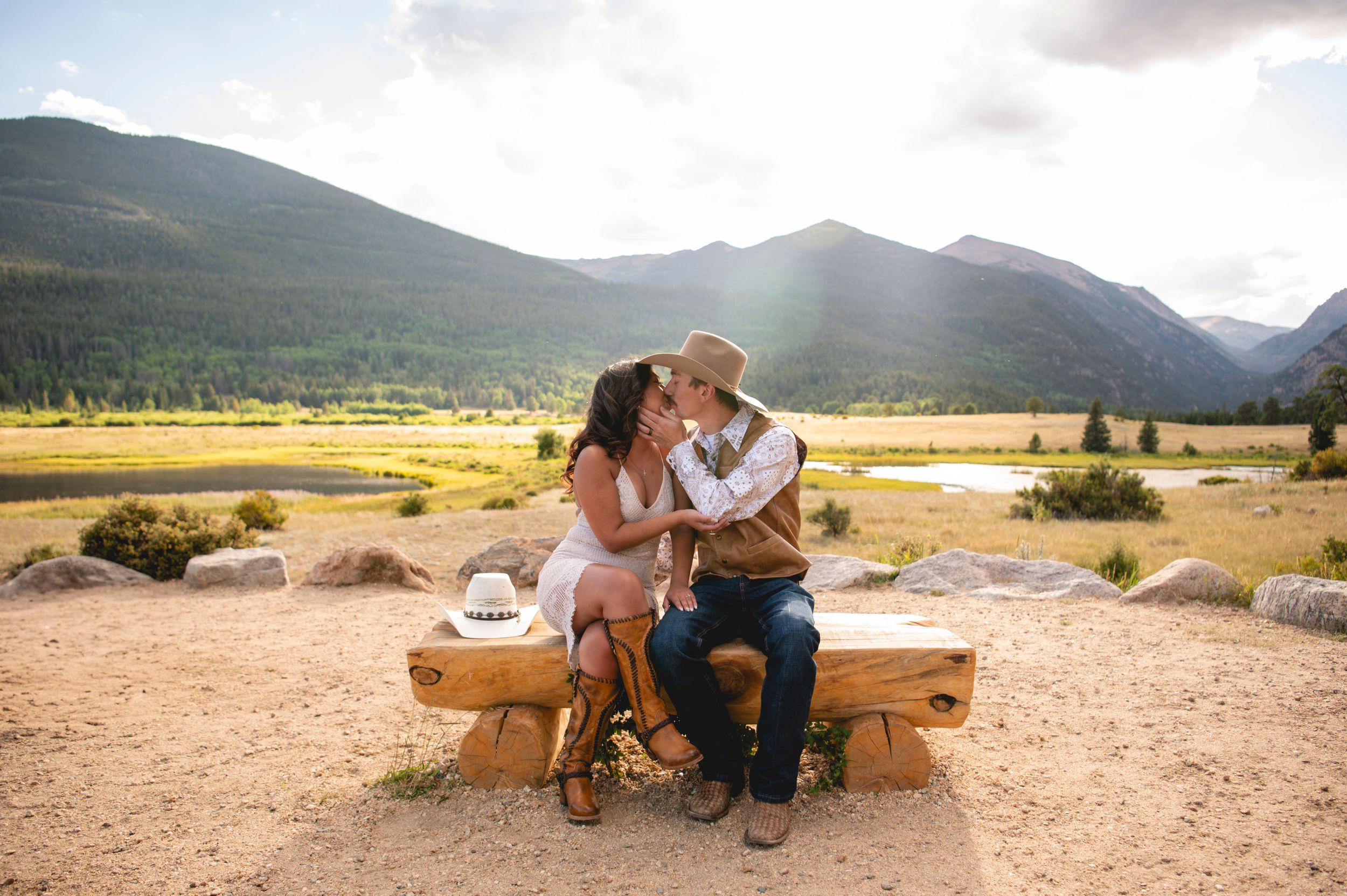 Couple sharing a romantic kiss on a rustic wooden bench with scenic mountain valley and golden fields in background.