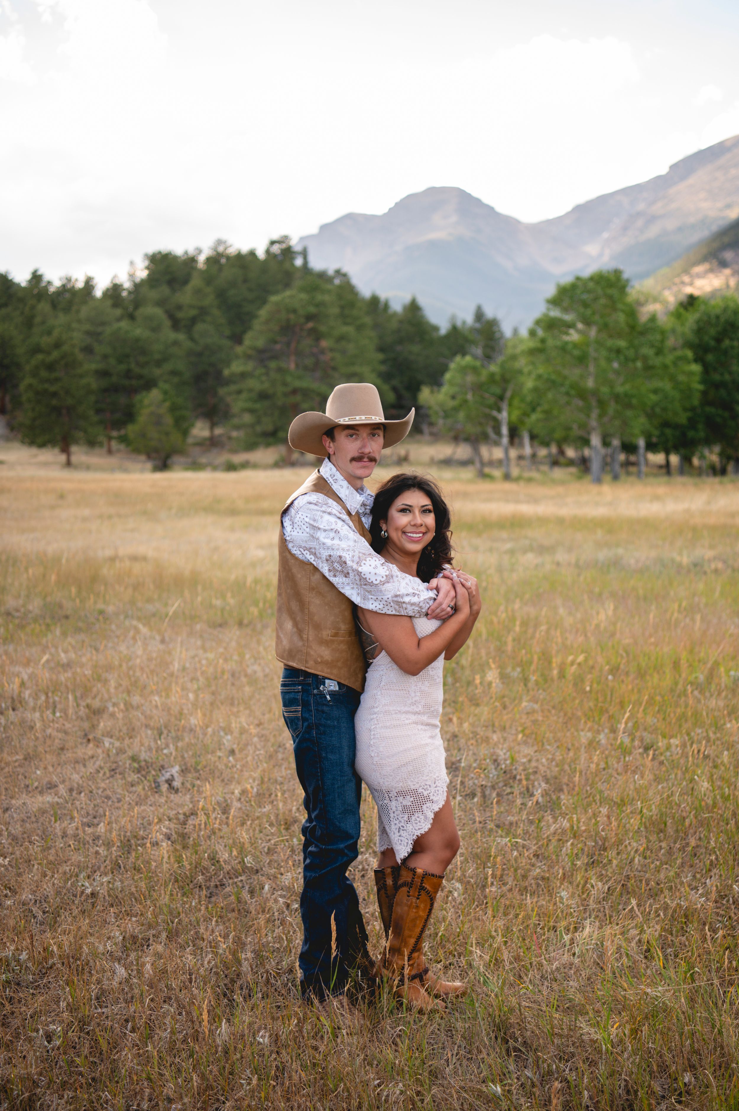 Couple embracing in golden meadow with mountain backdrop, woman in lace dress and boots, man in cowboy hat and vest.