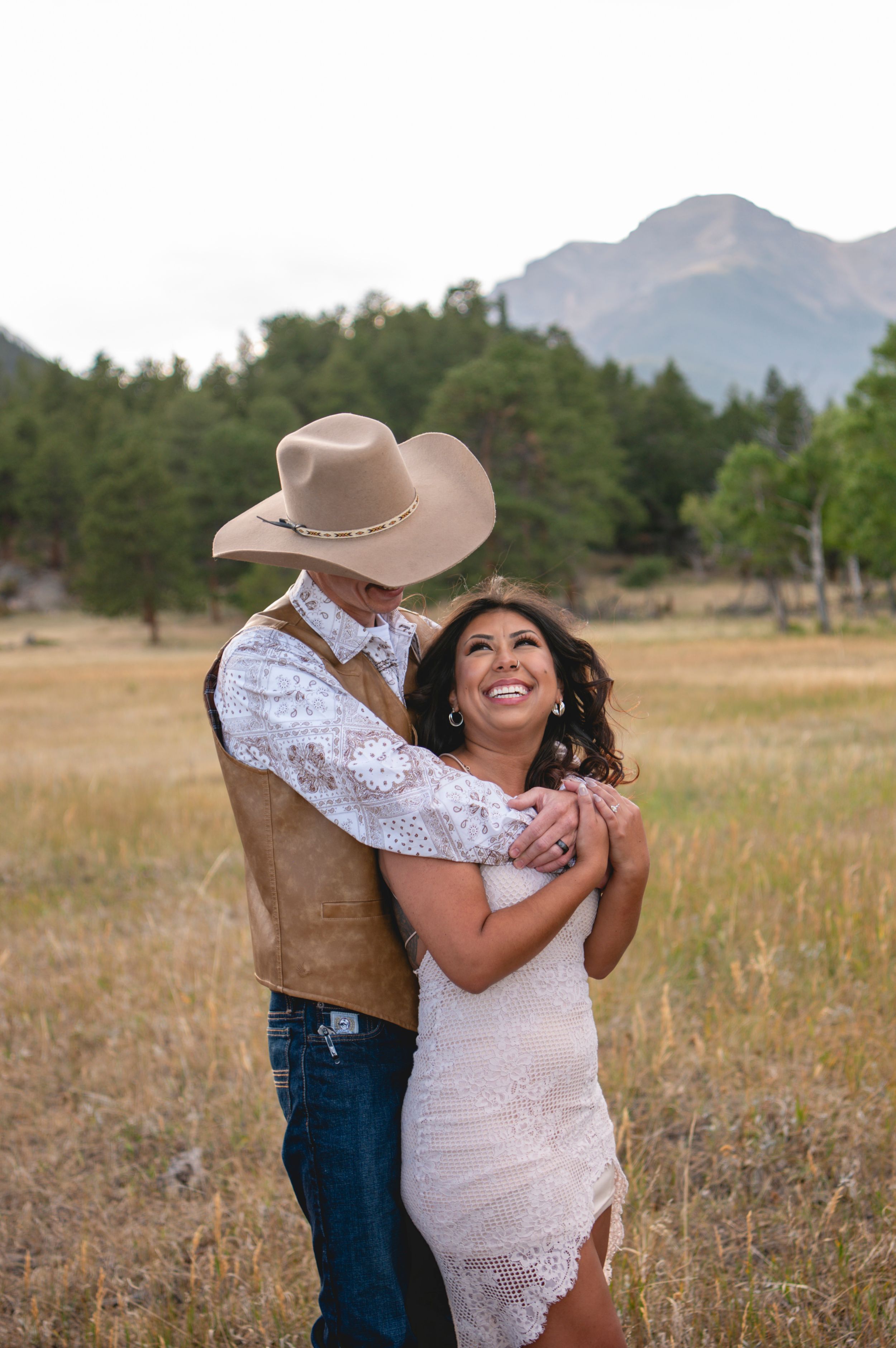 Couple embracing in a mountain meadow, woman in lace dress smiling, man in cowboy hat and vest, rustic western wedding.