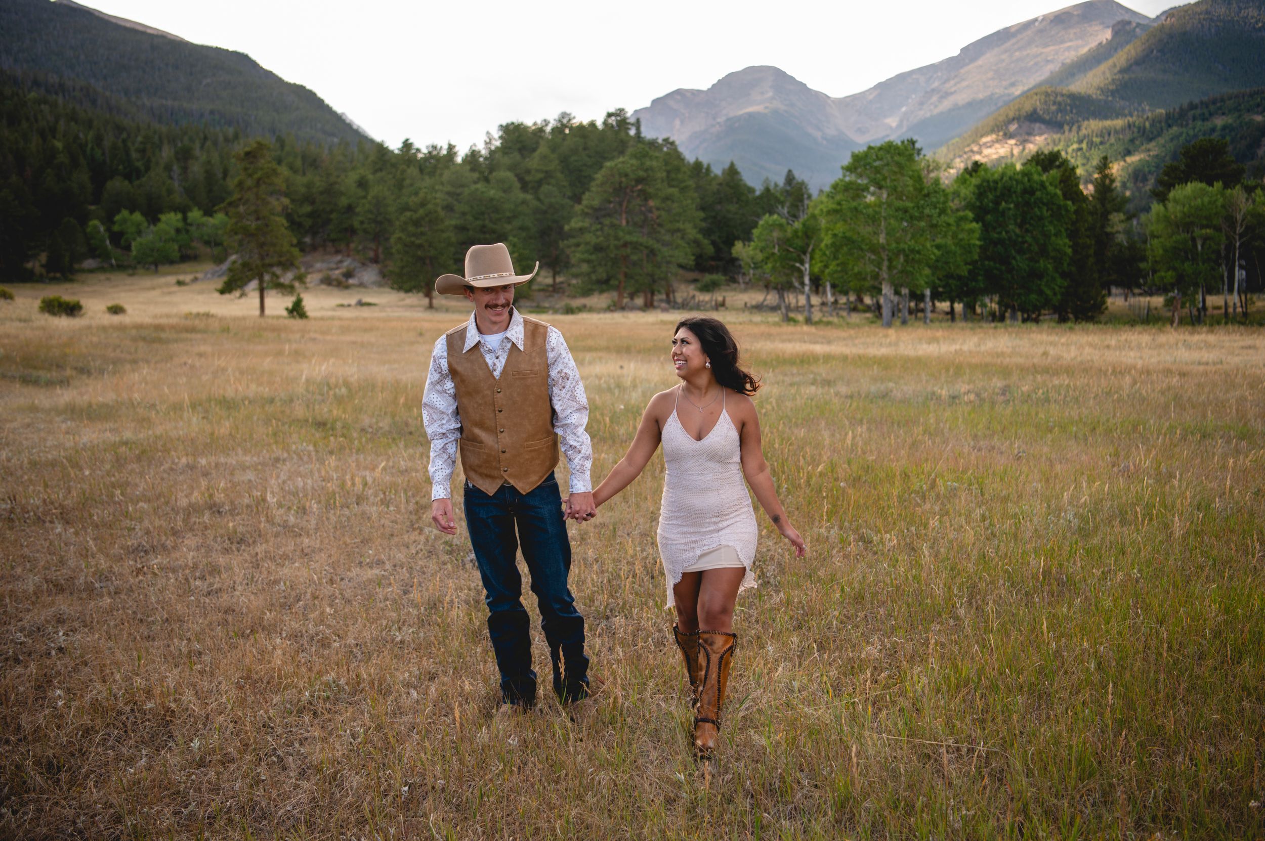 Couple in western attire holding hands in a golden meadow with mountain backdrop during outdoor wedding celebration.