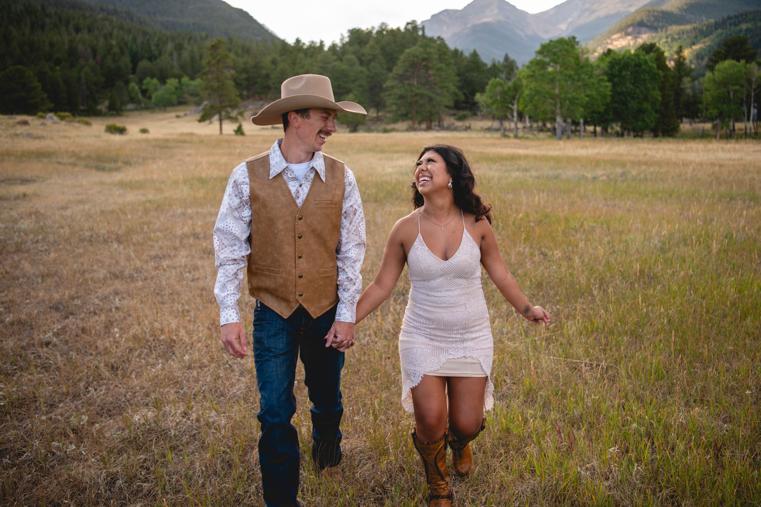 Couple holding hands walking in mountain meadow, man in cowboy hat and vest, woman in white dress and boots.
