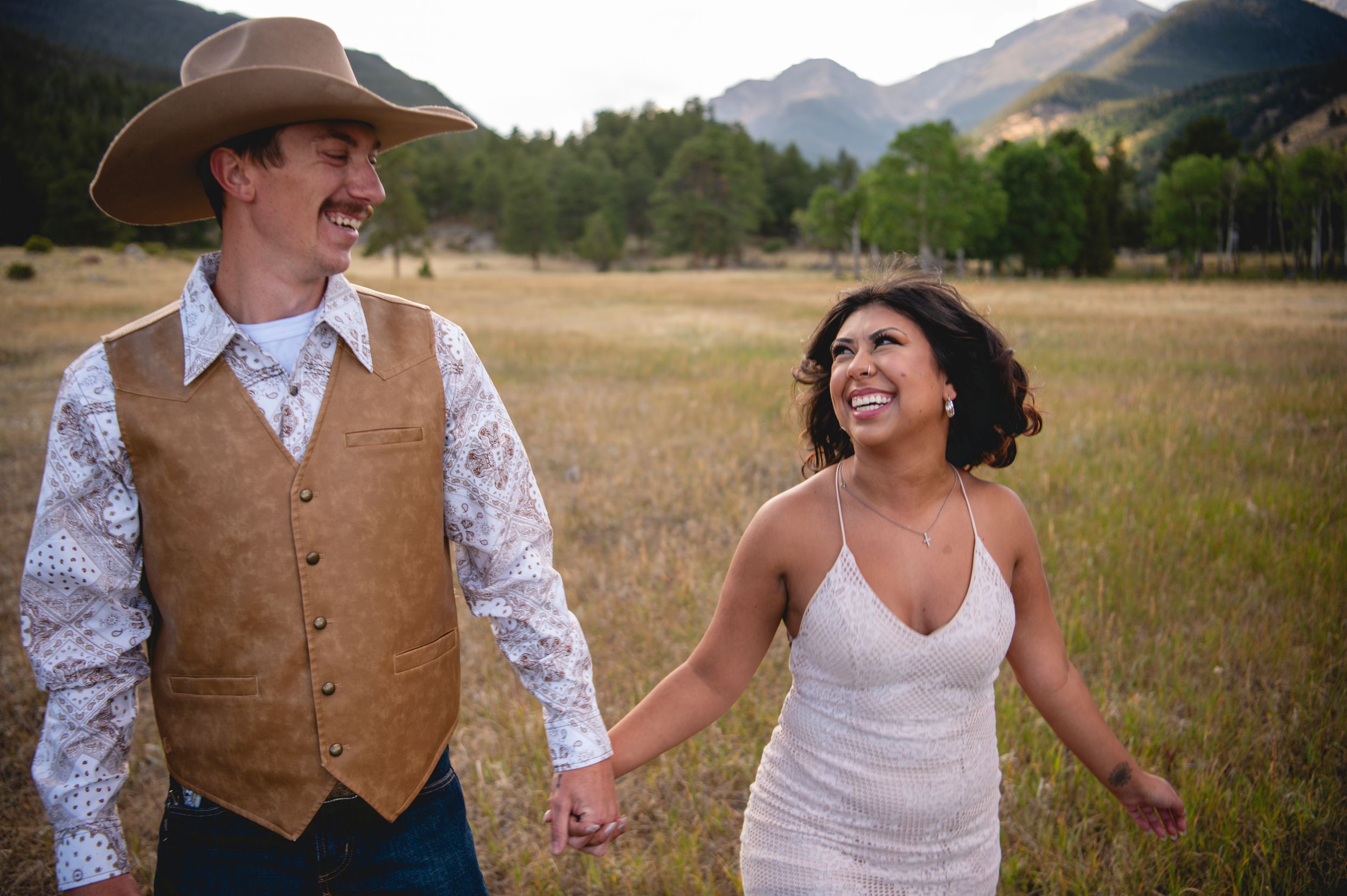 A joyful couple holds hands in a mountain meadow, the groom in a cowboy hat and vest, the bride in a white dress.