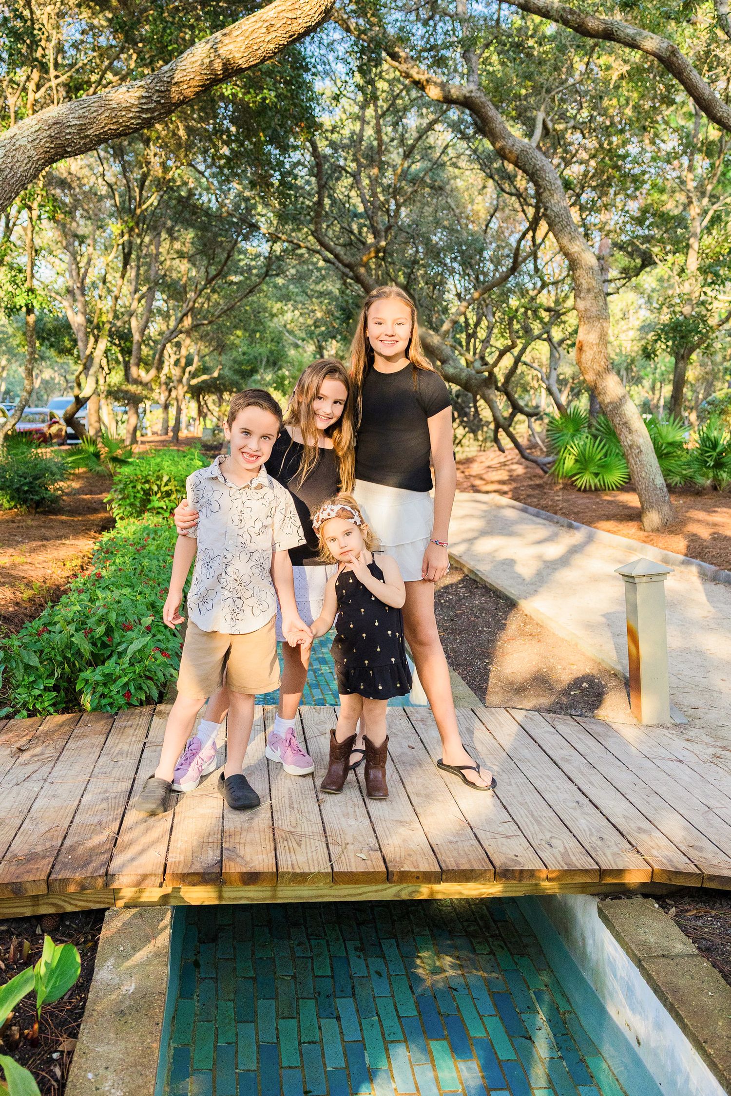 Four children smiling and posing on a wooden bridge surrounded by lush green trees in a park.