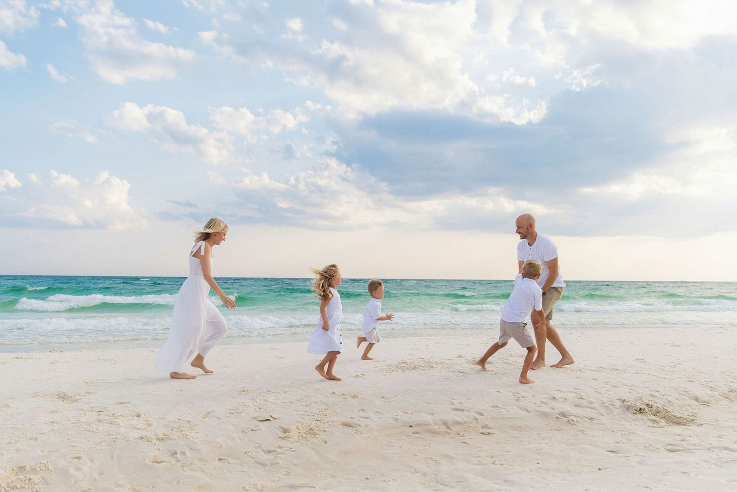 Family in white outfits joyfully walking on a sunny beach with turquoise ocean waves in the background.
