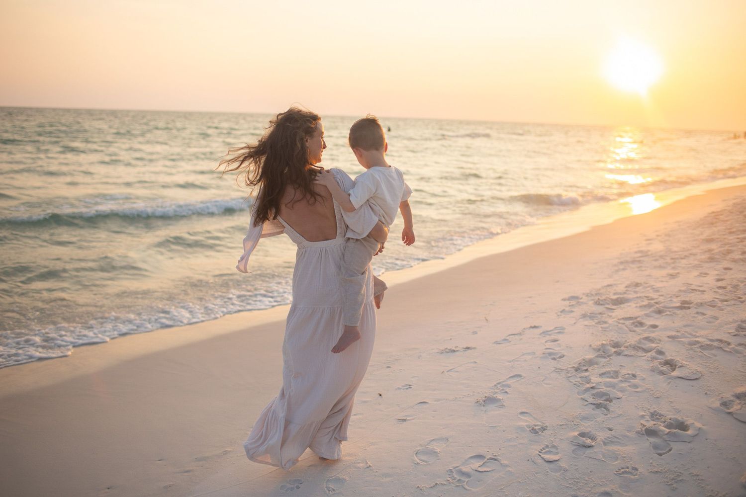 Woman in white dress carries child along sandy beach at golden sunset with ocean waves in background.