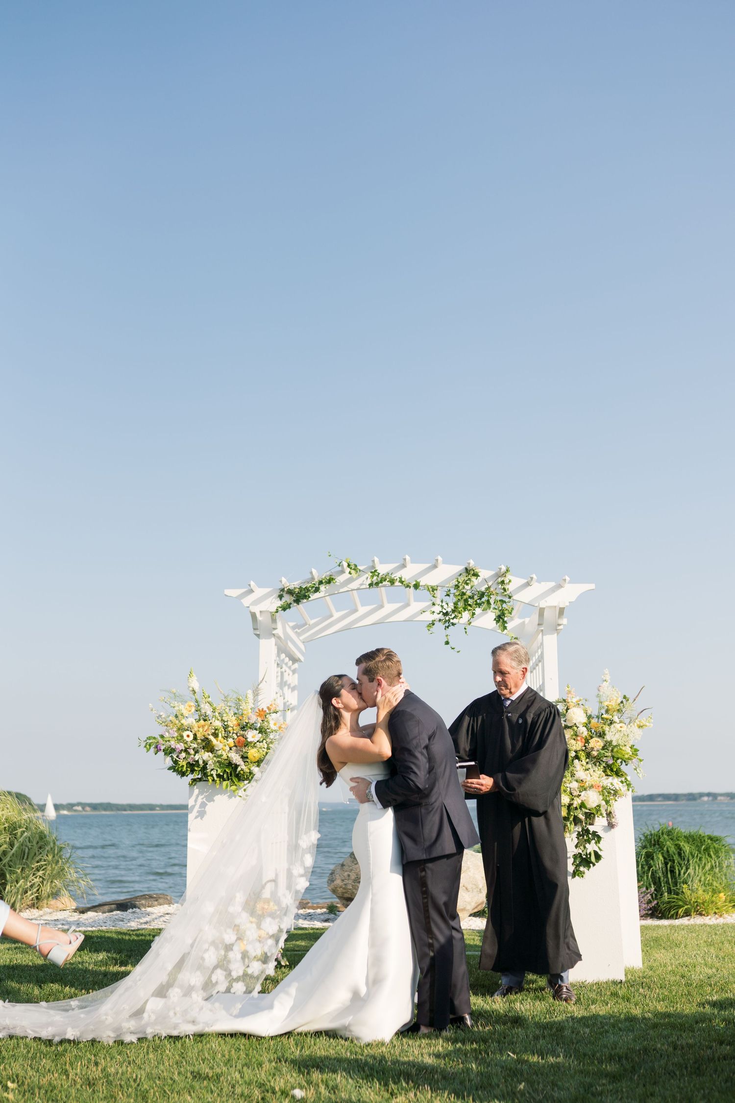 Bride and groom share first kiss at outdoor waterfront wedding ceremony under white floral arch on sunny day.