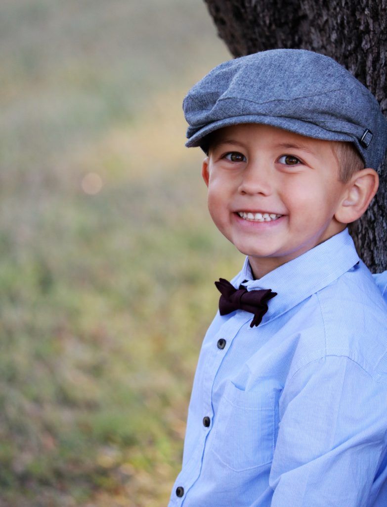 Smiling young boy wearing a flat cap and bow tie beside a tree