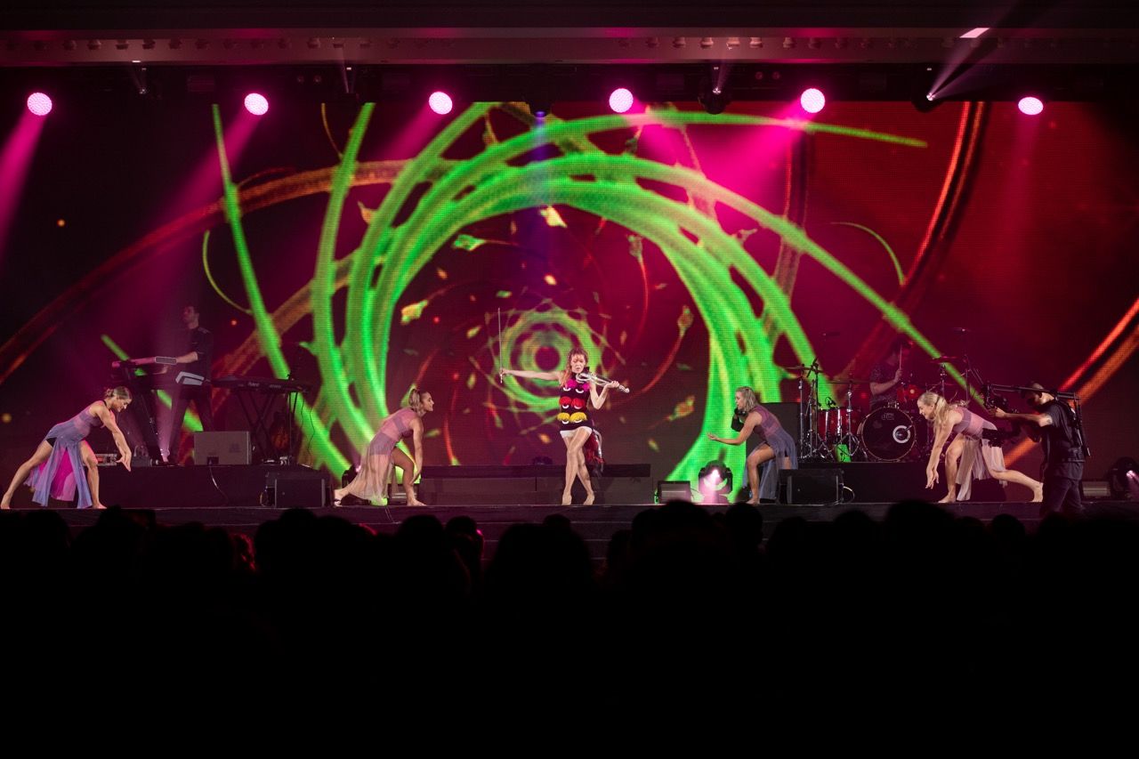 Performers on stage with vibrant green ribbon arcs and colorful lighting during a live theatrical show.