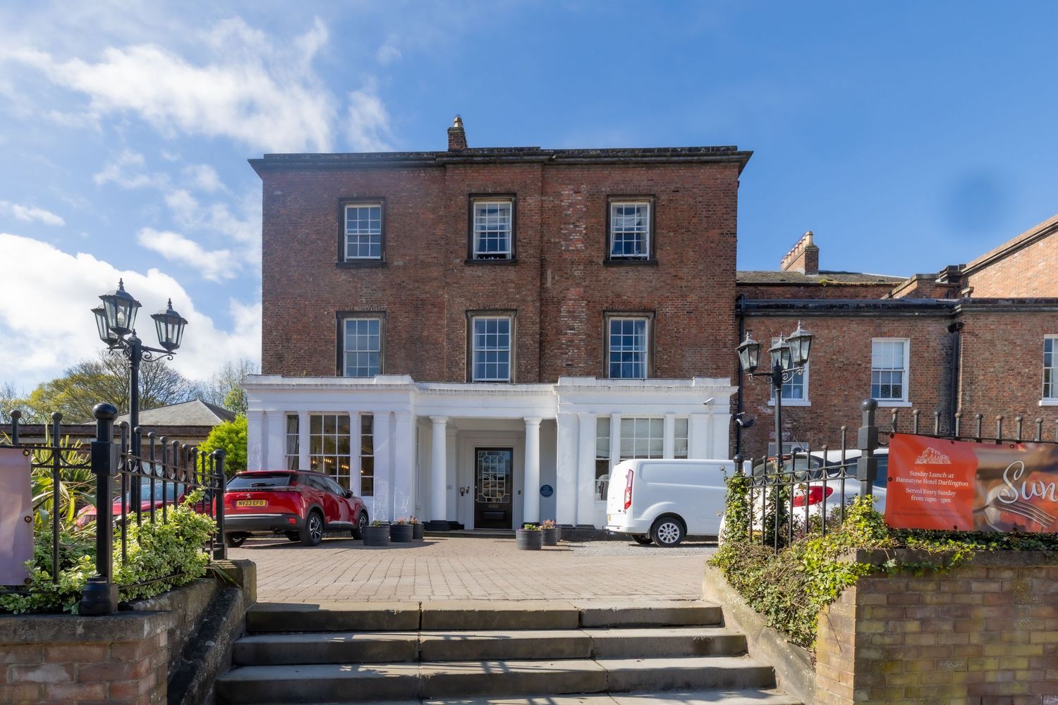 A grand red brick Georgian townhouse with white shopfront, stone steps, and parked vehicles on a sunny day. Bannatyne hotel, wedding