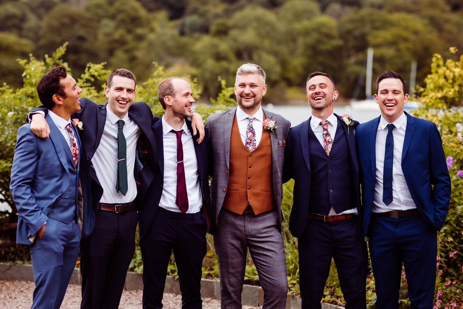 Three young ring bearers in navy suits and tan shoes sit on a dock, making funny faces at a wedding.