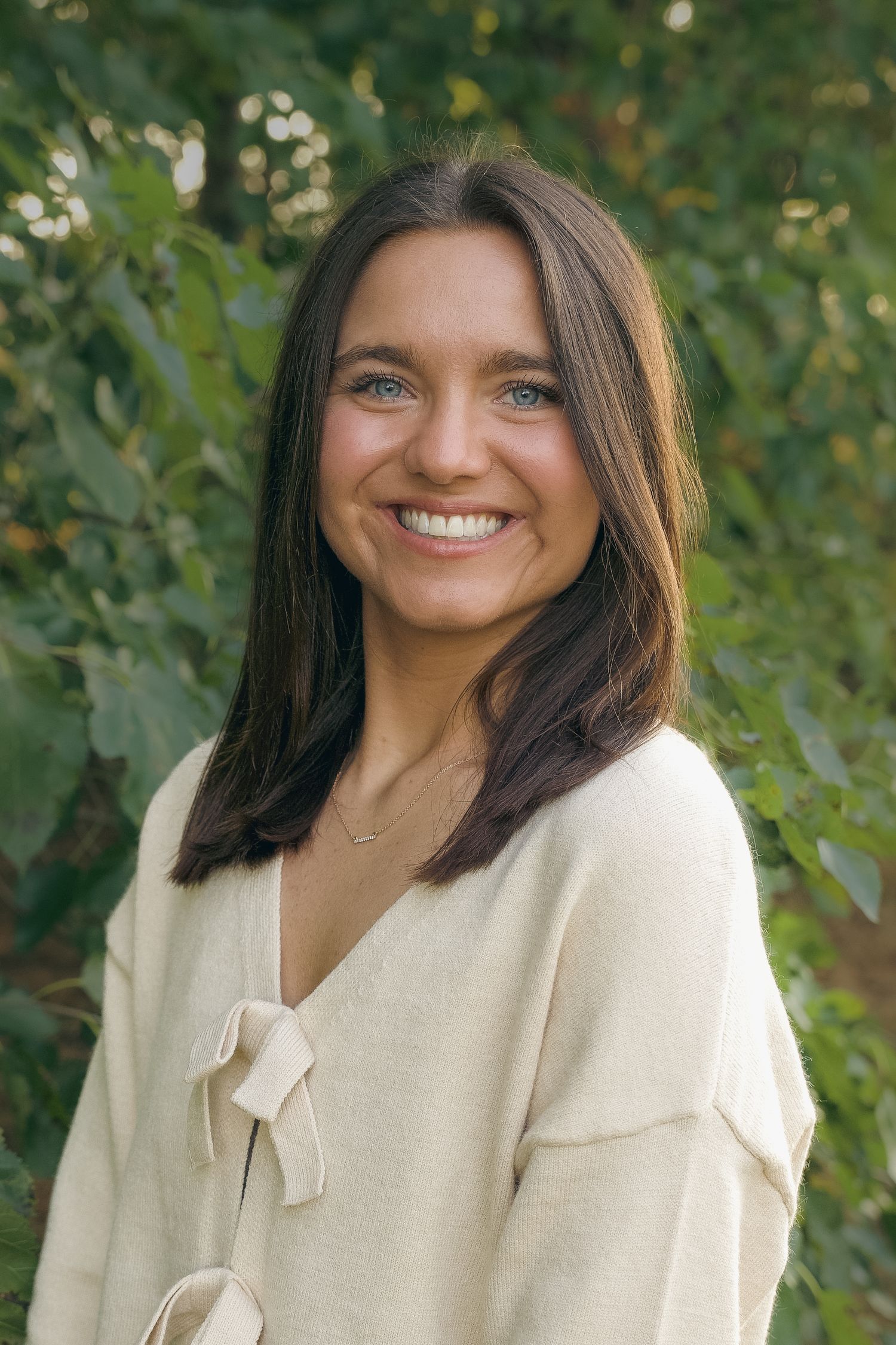 Smiling young woman with dark hair wearing a cream sweater, posing outdoors with green foliage background.