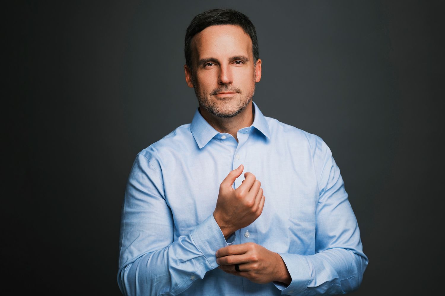 Professional man in light blue dress shirt adjusting cufflinks against dark studio background.