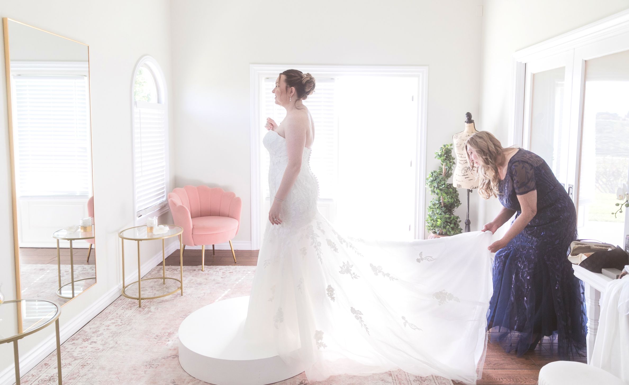 Bride in a white wedding gown being assisted with her train by a helper in a bright bridal suite.