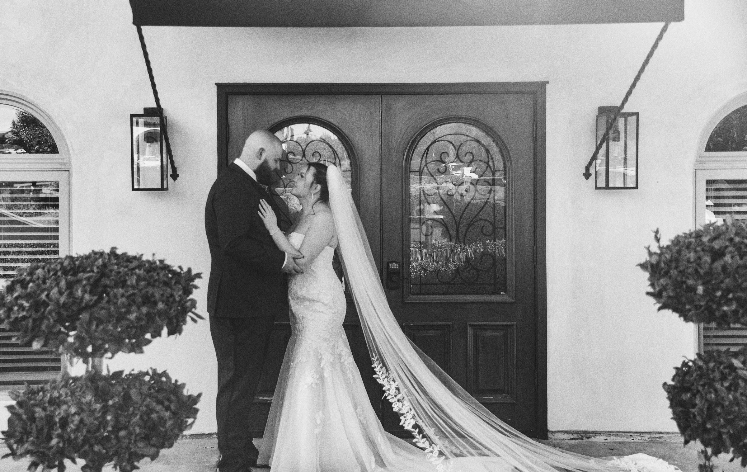 Black and white wedding photo of bride and groom embracing before ornate door with topiary bushes.