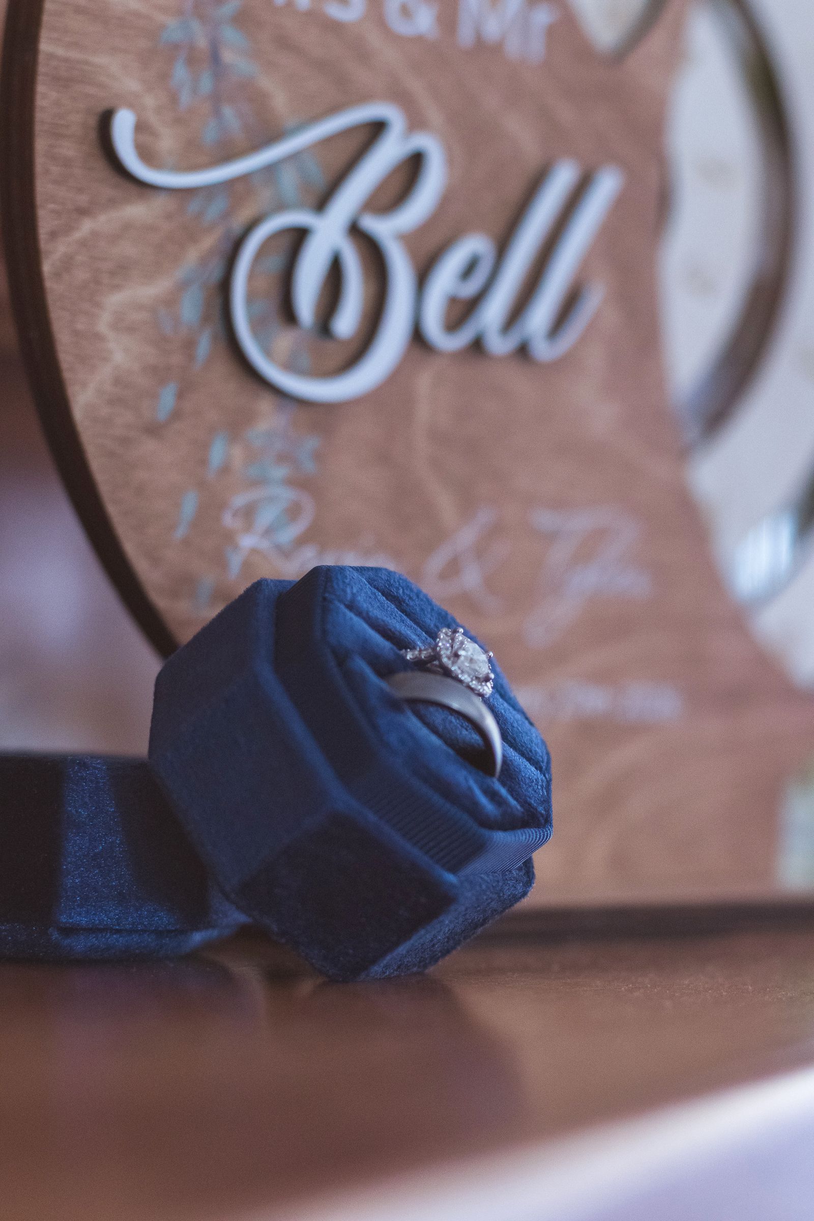 Diamond engagement ring displayed in a navy blue velvet box with a Bell wedding sign in the background.