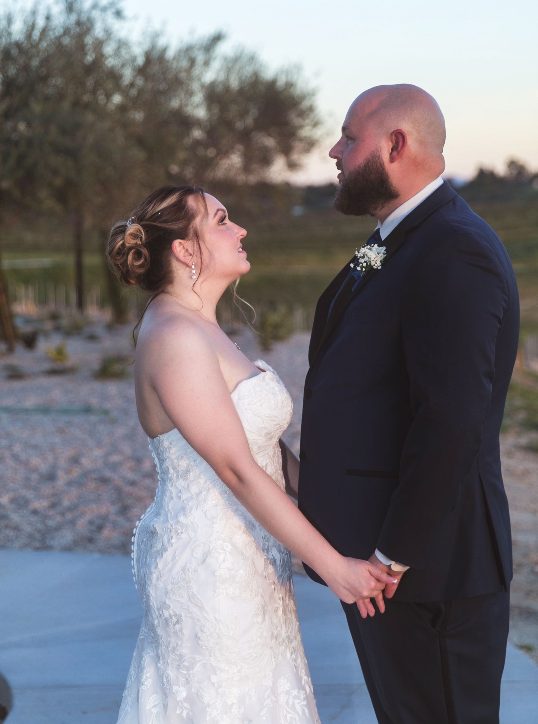 Bride in white lace gown and groom in navy suit hold hands and gaze at each other during outdoor wedding ceremony at sunset.