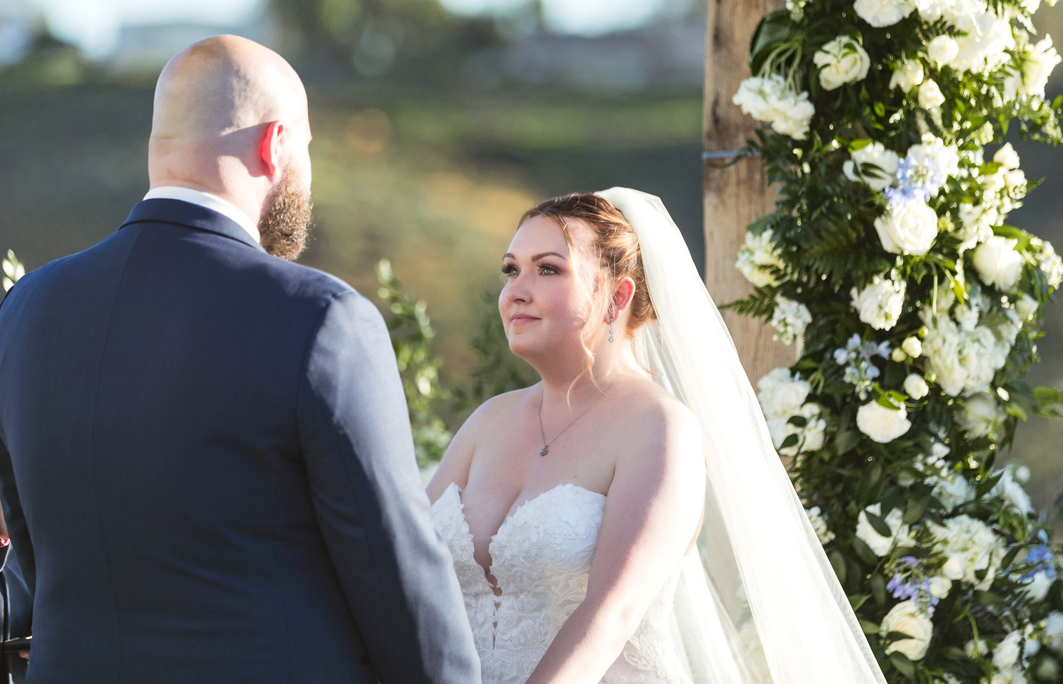 Bride in white gown and veil smiles at groom during outdoor wedding ceremony with floral arch backdrop.