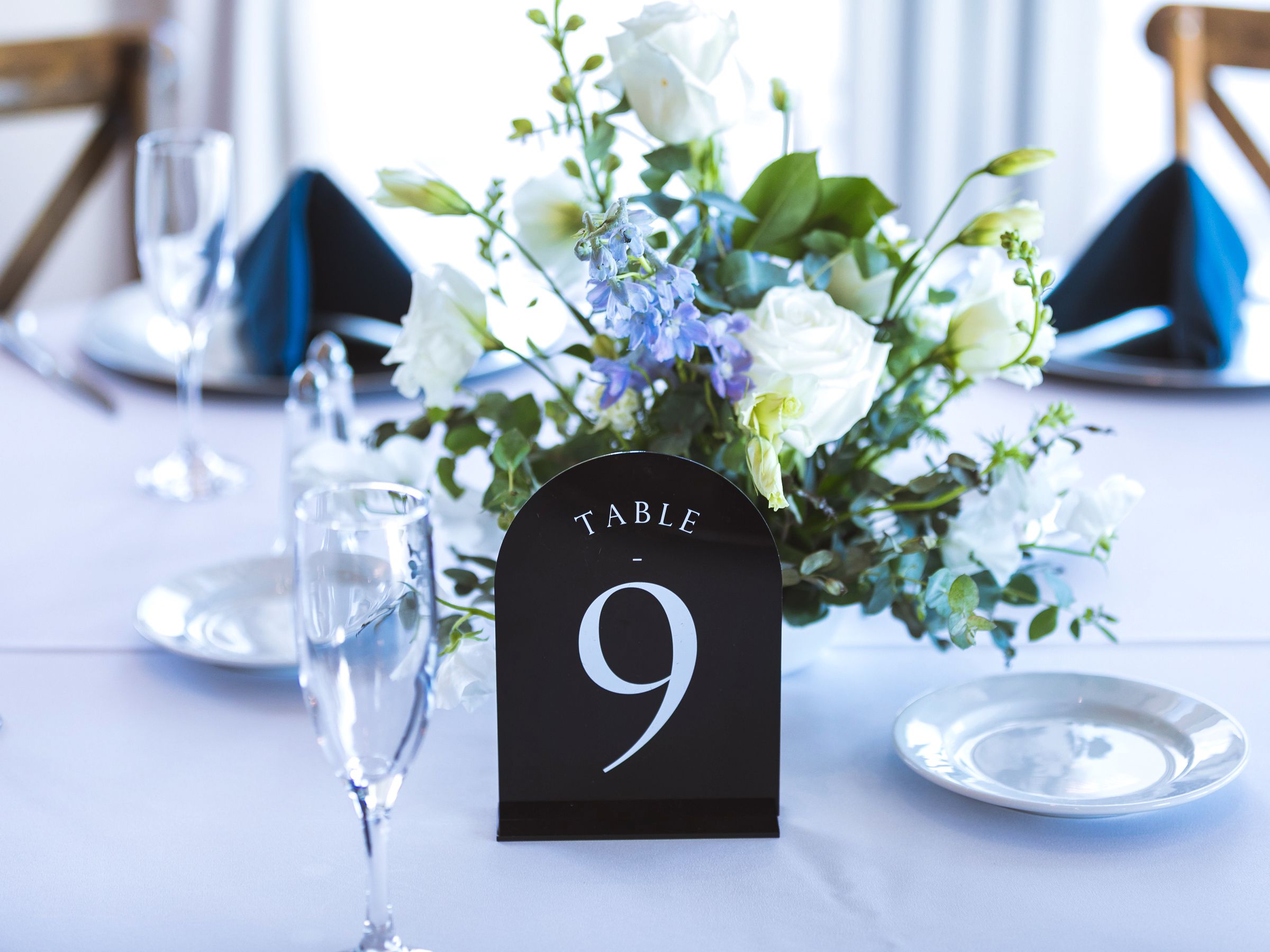 Elegant wedding table setting with a black table number 9 sign, white floral centerpiece, and blue napkins.