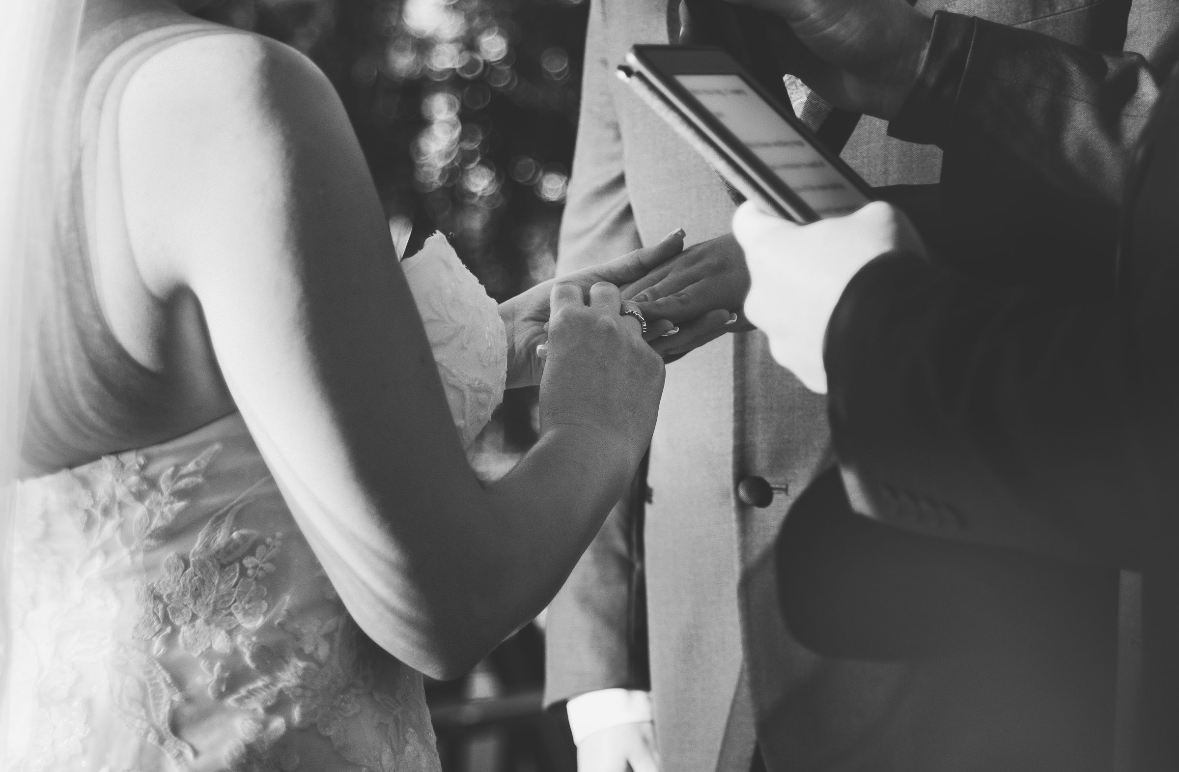Black and white wedding ceremony photo of couple exchanging rings with officiant holding tablet.