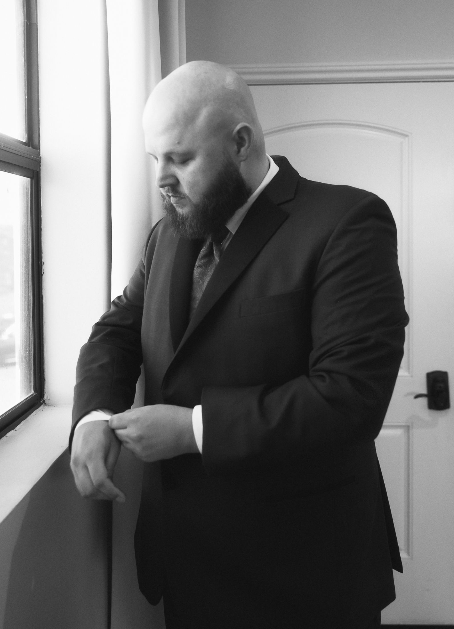 Bald bearded man in black suit adjusting cufflinks by window, black and white professional portrait photo.