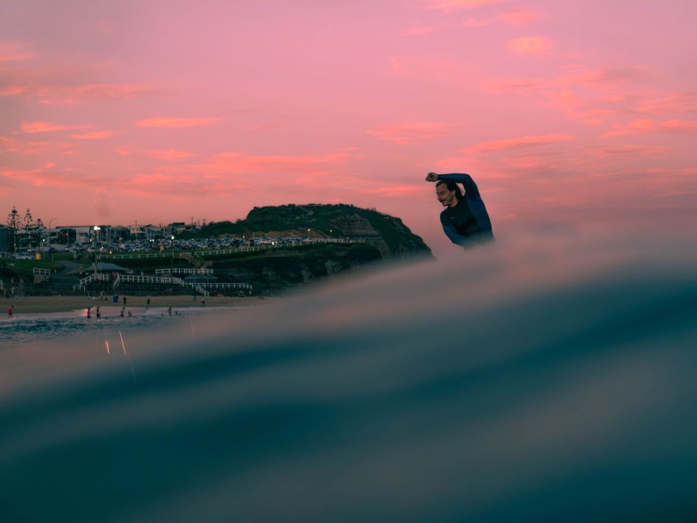 photo of a surfer with pink sky and city lights at sunset behind a wave at Bae Beach, Newcastle NSW