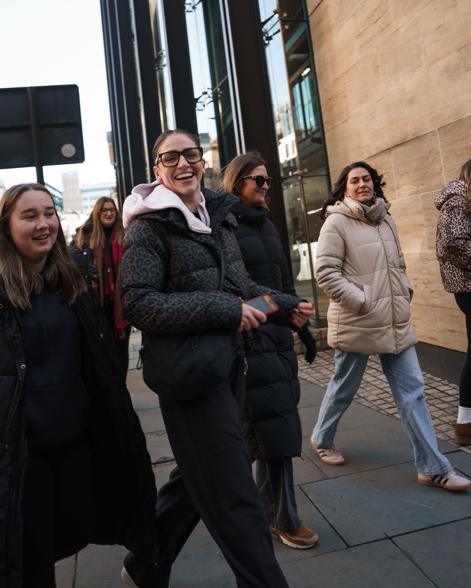 Group of smiling women walking outdoors in winter coats on a city sidewalk.