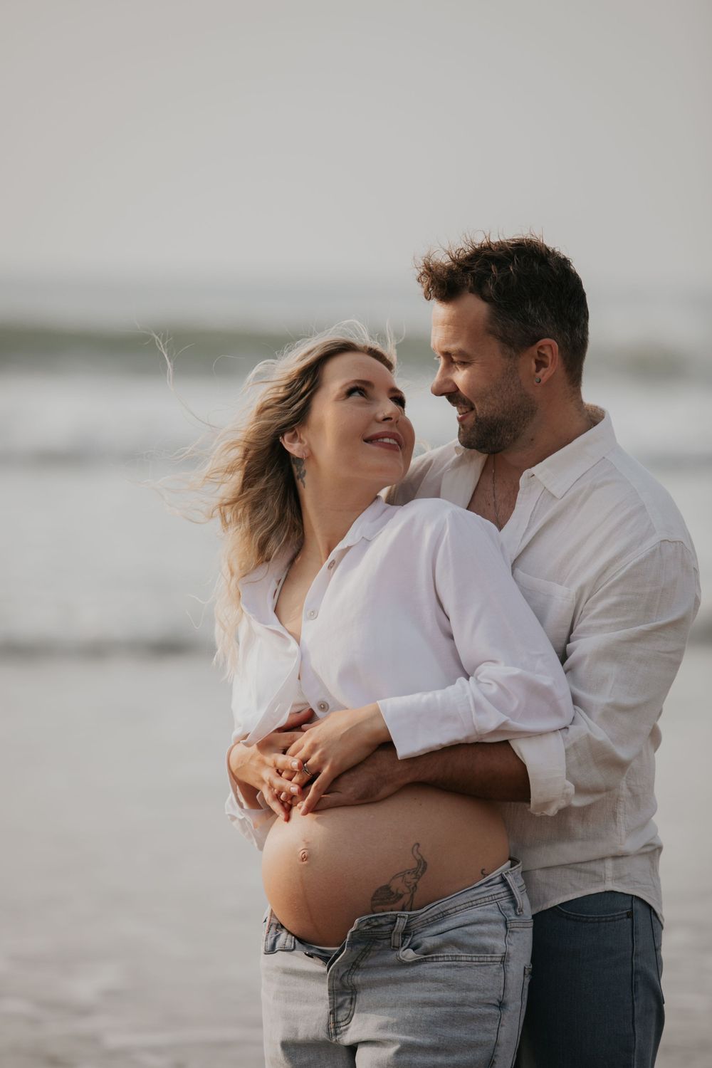 Couple embracing on beach, man holding pregnant woman's baby bump while smiling at each other. Cornwall Maternity Photographer